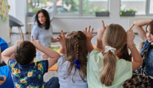 A Happy Female Teacher Sitting And Playing Hand Games With A Group Of Little Schoolchildren